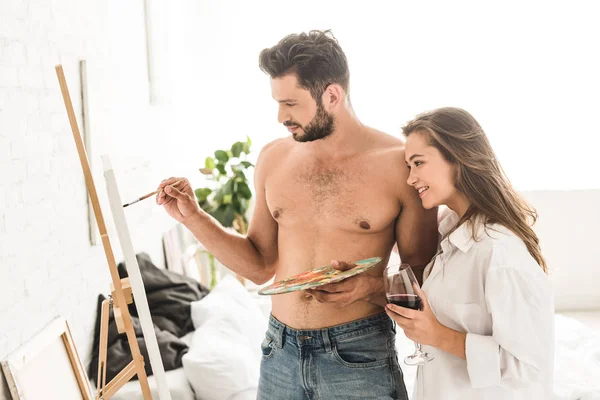 Sexy jeune homme dessin tandis que la fille debout avec du vin et en regardant la peinture — Photo de stock