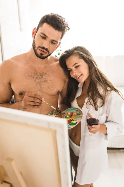 Selective focus of sexy young man drawing while girl standing with wine and looking at painting — Stock Photo