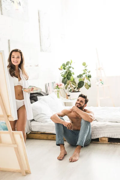 Повнометражний вигляд дівчини, що малює пензлем і чоловіком, що сидить на підлозі біля ліжка і посміхається, дивлячись на картину — стокове фото