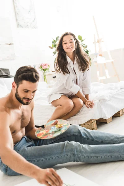 Hombre dibujo mientras está sentado en el suelo y la niña sonriendo mientras está sentado en la cama - foto de stock