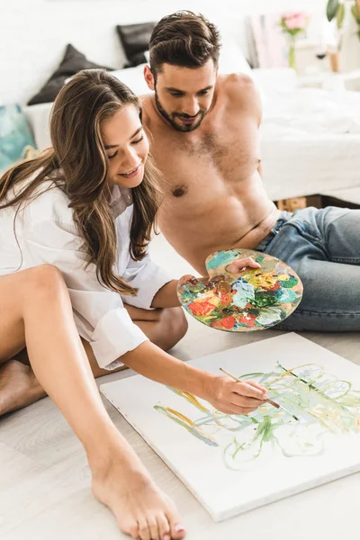 Joven chica dibujo con pincel y pinturas de colores, mientras que el hombre sin camisa sentado junto a su novia - foto de stock