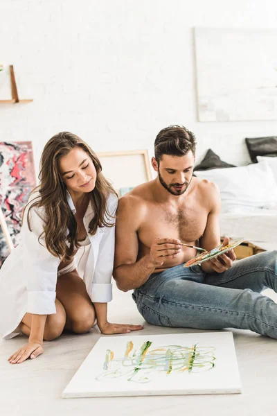 Sexy shirtless man drawing while girl sitting behind guy, smiling and looking at painting — Stock Photo