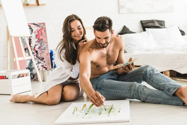 Sexy pareja de artistas sentado en el suelo y sonriendo mientras el hombre dibujo con cepillo - foto de stock