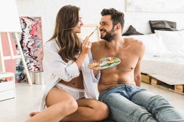 Feliz pareja sentada en el suelo y mirándose mientras chica tratando de pintar la cara de chico - foto de stock