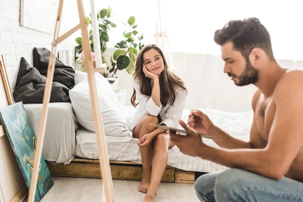 Selective focus of sexy shirtless man drawing and girl smiling while sitting in bed and looking at guy — Stock Photo