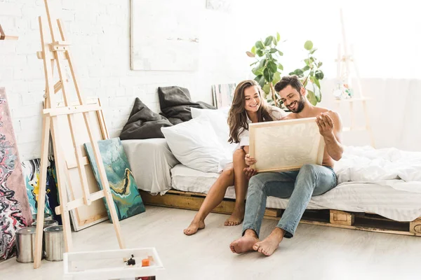 Feliz sexy pareja sentado en la cama mientras hombre sosteniendo pintura y mostrando a chica - foto de stock