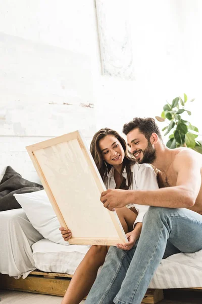 Joven pareja sentado en cama y sonriendo mientras hombre sosteniendo pintura y chica mirando chico - foto de stock