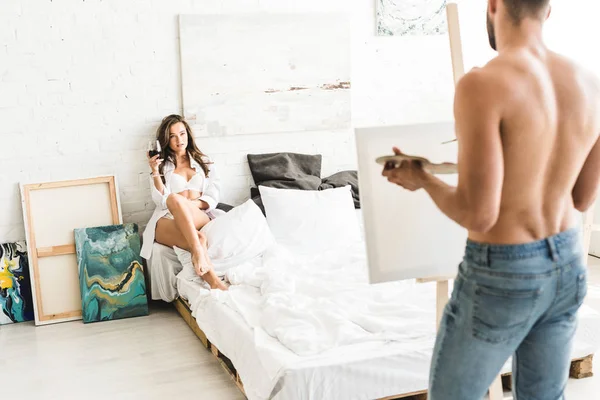 Enfoque selectivo de la chica sentada en la cama con vino y posando para el hombre sin camisa dibujo novia - foto de stock