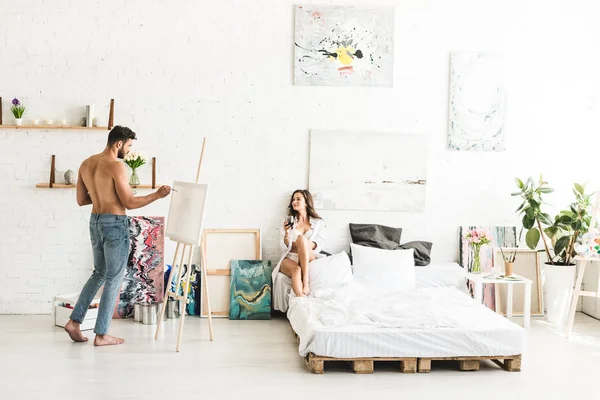 Вид в полный рост на дерзкого мужчину, рисующего подругу с цветными пайетками, пока девушка сидит в постели с вином — стоковое фото