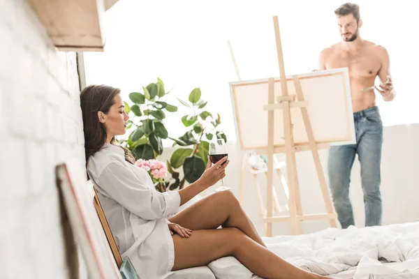 Vue latérale de la fille assise dans le lit avec du vin tandis que l'homme torse nu dessin petite amie avec brosse et peintures colorées — Photo de stock