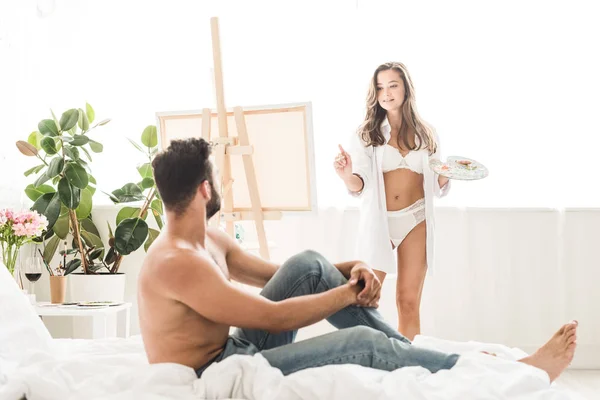 Sexy girl in white underwear drawing man while guy sitting in bed — Stock Photo