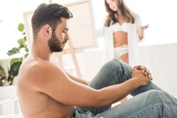 Selective focus of sexy shirtless man sitting with closed eyes in bed while girl drawing guy — Stock Photo