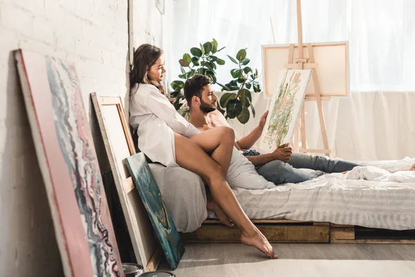 Side view of sexy young couple looking at painting while man lying in bed and girl sitting behind guy — Stock Photo