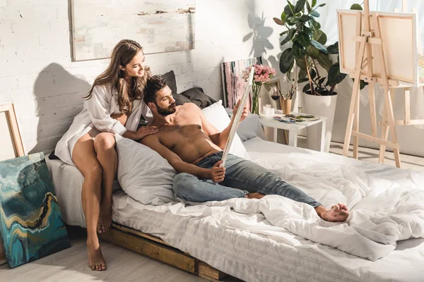 Sexy joven pareja sonriendo y mirando a la pintura mientras hombre acostado en cama y chica sentado detrás de chico - foto de stock
