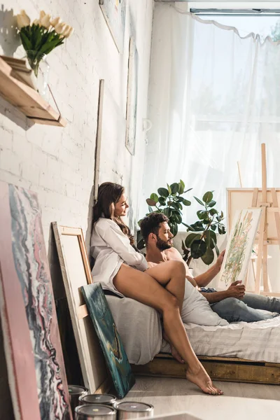 Side view of sexy young couple looking at painting while man lying in bed and girl sitting next to guy — Stock Photo