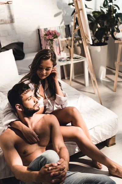 Sexy young man sitting on floor near bed while girl looking at guy and sitting behind boyfrined — Stock Photo