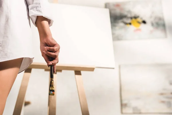 Cropped view of girl holding paintbrush near easel — Stock Photo