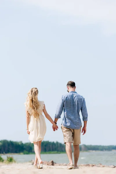 Вид ззаду дорослої пари, що йде вздовж пляжу і тримає руки в сонячний день — стокове фото