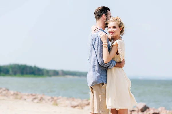 Mujer rubia sonriente abrazando novio barbudo en la playa - foto de stock