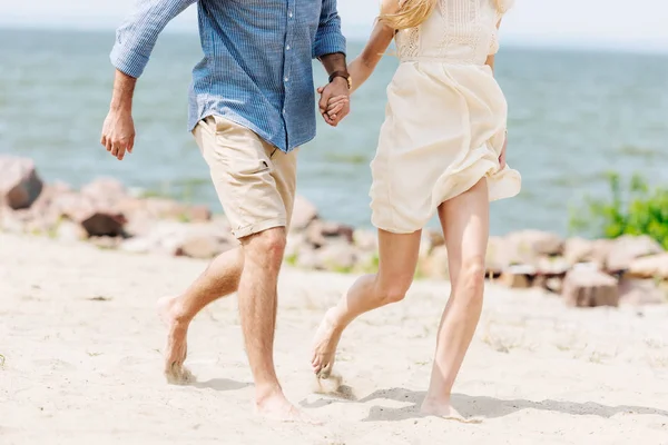 Vue recadrée du jeune couple pieds nus tenant la main tout en courant le long de la plage — Photo de stock