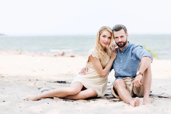 Beautiful young barefoot couple sitting on blanket at beach near sea — Stock Photo