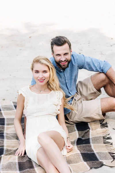Hermosa y sonriente joven pareja sentada en manta a cuadros en la playa de arena - foto de stock