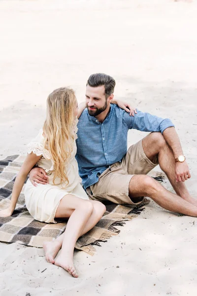 Hermosa pareja joven descalza sentado en manta y mirándose en la playa - foto de stock