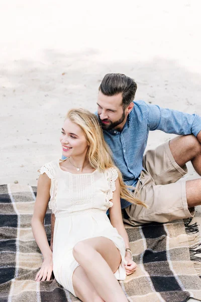 Smiling beautiful young couple sitting on plaid blanket at sandy beach — Stock Photo