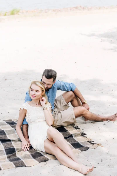 Jovem casal descalço sentado em cobertor e olhando para longe na praia perto do mar — Fotografia de Stock