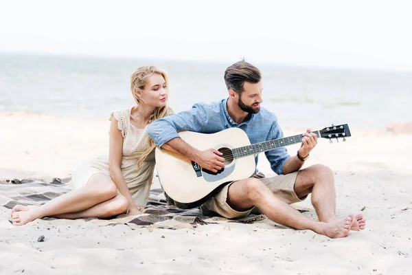 Hermosa joven descalza mujer sentada en manta con novio tocando la guitarra acústica en la playa cerca del mar - foto de stock