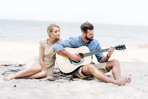 Schöne junge barfüßige Frau auf Decke sitzend mit Freund, der Akustikgitarre am Strand am Meer spielt — Stockfoto