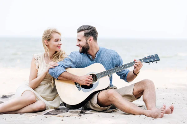 Hermosa joven sentada en manta y mirando novio tocando la guitarra acústica en la playa cerca del mar - foto de stock