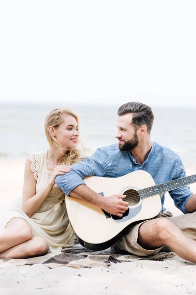 Sorrindo bela jovem descalça mulher sentada no cobertor e olhando para o namorado com guitarra acústica na praia perto do mar — Fotografia de Stock