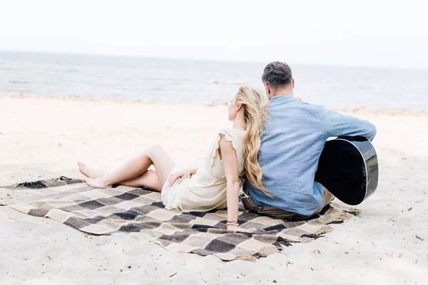 Back view of young blonde barefoot woman sitting on checkered blanket near boyfriend with acoustic guitar at beach near sea — Stock Photo