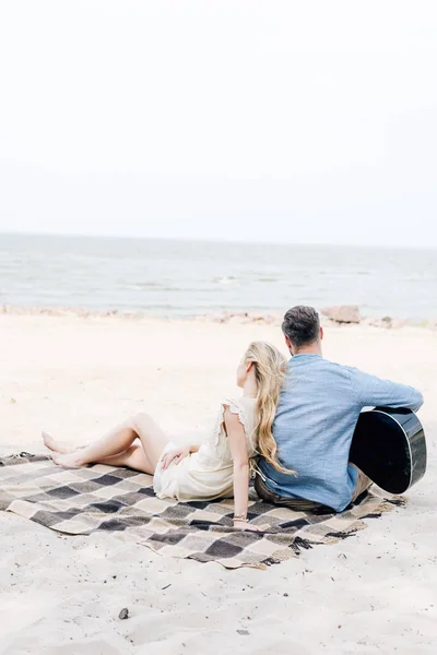 Vue arrière de la jeune femme blonde pieds nus assise sur une couverture à carreaux près du petit ami avec guitare acoustique à la plage près de la mer — Photo de stock
