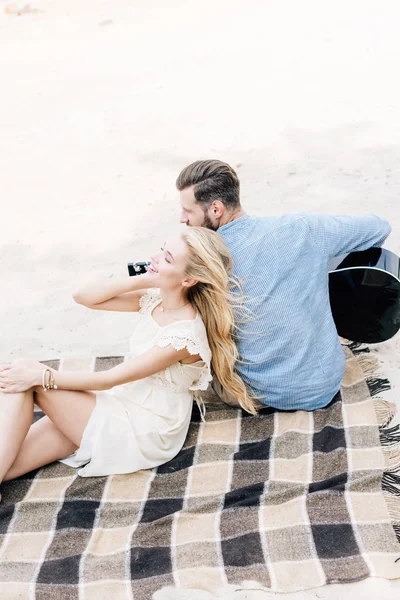 Heureuse jeune femme blonde assise sur une couverture à carreaux près du petit ami avec guitare acoustique à la plage de sable — Photo de stock