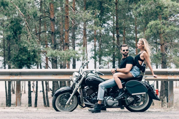 Side view of young sexy couple of bikers on black motorcycle on road near green forest — Stock Photo