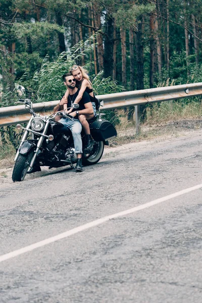 Молодая пара байкеров обнимается на черном мотоцикле на дороге возле зеленого леса — стоковое фото