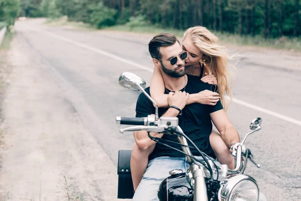 Joven tierna pareja de ciclistas abrazándose en motocicleta negro en la carretera cerca de bosque verde - foto de stock