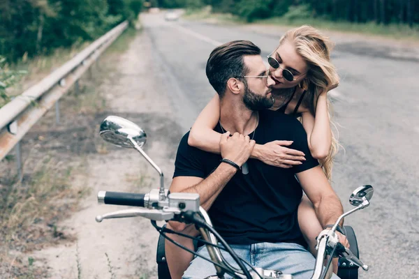 Jovem casal de motociclistas abraçando e olhando uns para os outros na motocicleta preta na estrada perto de floresta verde — Fotografia de Stock