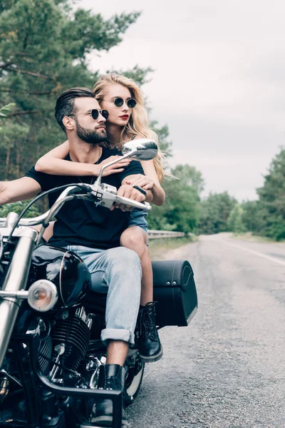 Jovem casal de motociclistas abraçando e olhando para longe na motocicleta preta na estrada perto de floresta verde — Fotografia de Stock