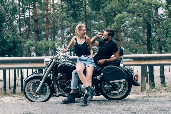 Young happy couple of bikers on black motorcycle on road near green forest — Stock Photo