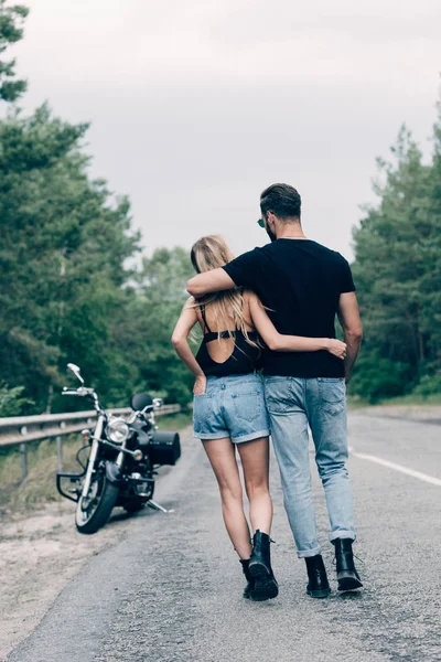 Back view of young couple of bikers walking along road and embracing near black motorcycle — Stock Photo