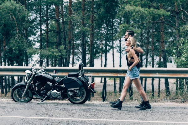 Side view of young couple of bikers walking along road and embracing near black motorcycle and green forest — Stock Photo