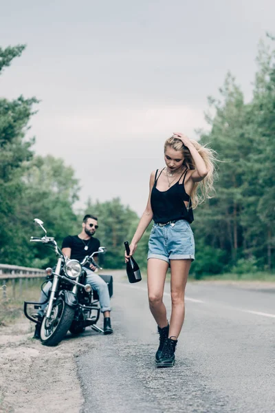 Selective focus of young woman walking on road with bottle of alcohol near boyfriend on black motorcycle — Stock Photo