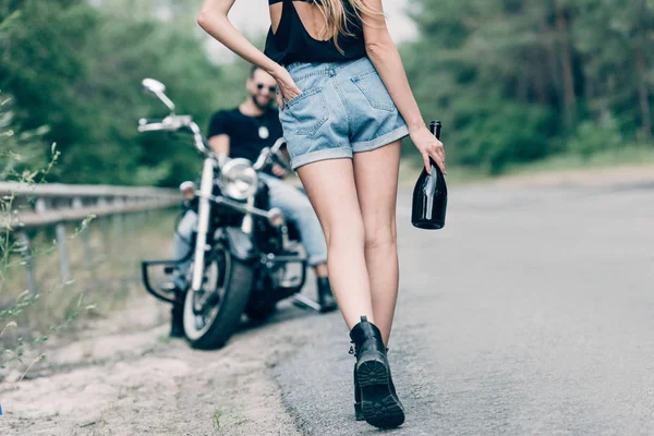 Частковий вид на молоду жінку, що йде по дорозі з пляшкою алкоголю біля хлопця на чорному мотоциклі — стокове фото
