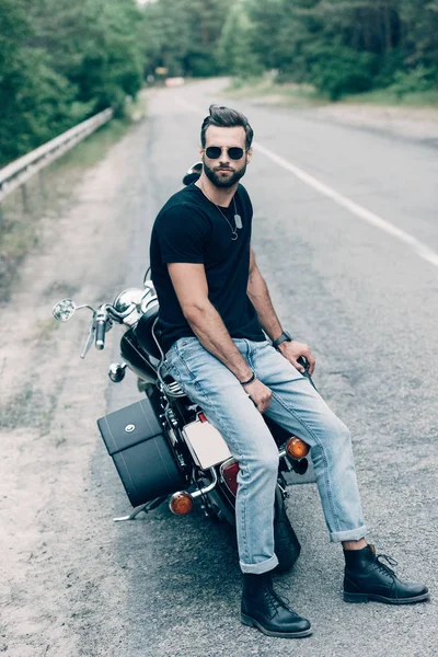 Красивый мотоциклист в солнечных очках сидит на черном мотоцикле на дороге возле зеленого леса — стоковое фото