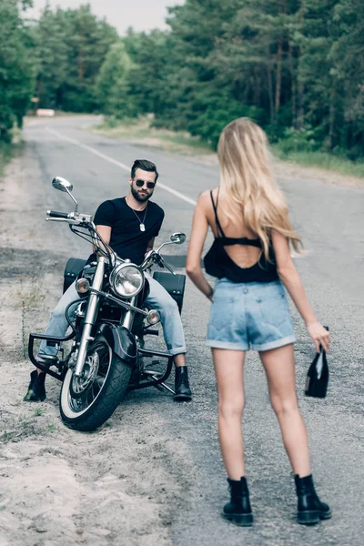 Vista trasera de la chica caminando por la carretera con botella de alcohol cerca de novio guapo en gafas de sol en motocicleta negro - foto de stock