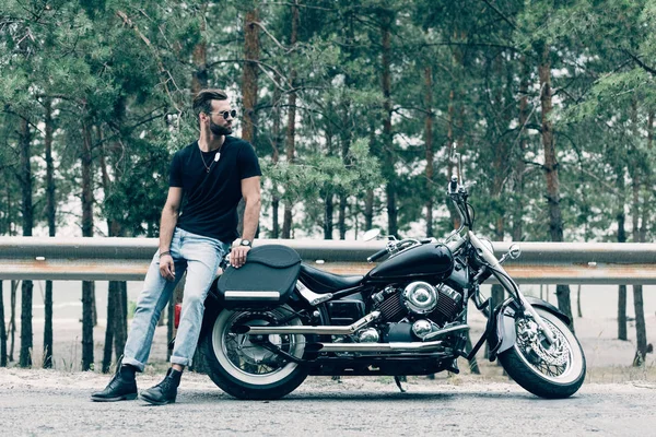 Schöner Motorradfahrer mit Sonnenbrille steht neben schwarzem Motorrad auf Straße nahe grünem Wald — Stockfoto