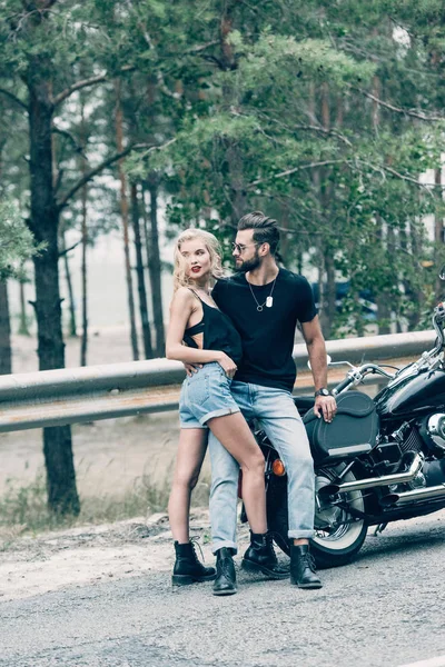 Молодая сексуальная пара мотоциклистов обнимаются и глядя в сторону возле черного мотоцикла на дороге возле зеленого леса — стоковое фото